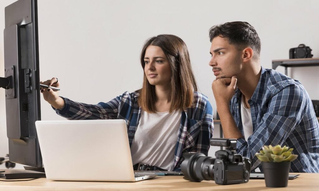 Frau erklärt Kollegen etwas am PC: Ihre Webseiten auf SEO zu optimieren, ist für Versicherer wichtig, damit Kunden auf Produkte und Dienstleistungen aufmerksam werden.