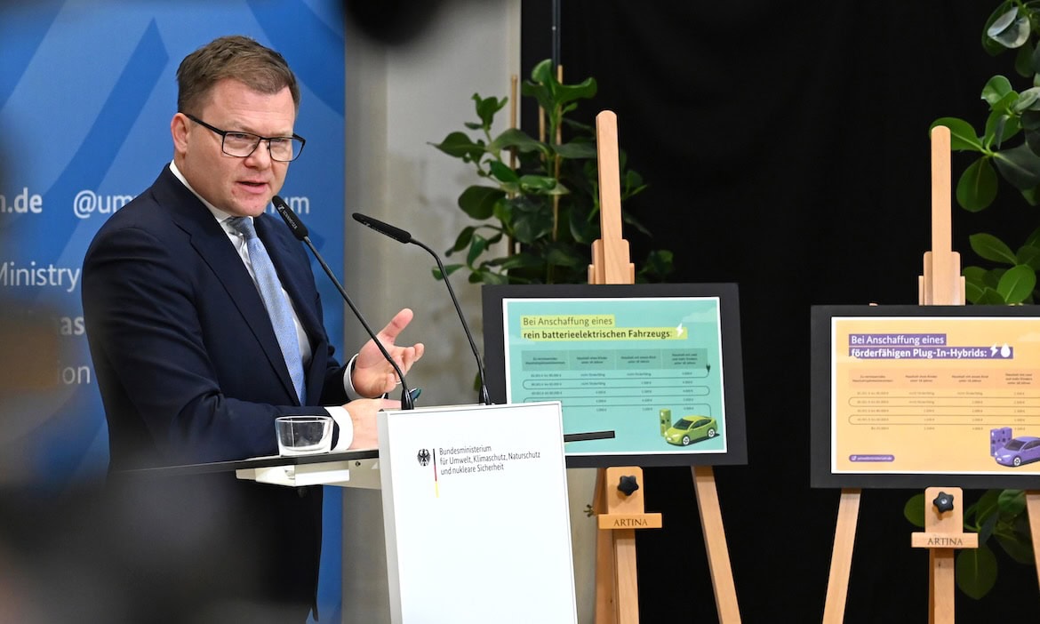 Mann bei Präsentation über E-Auto-Förderung, Infotafeln im Hintergrund.