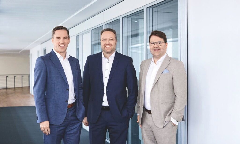 Männer in Business-Anzügen vor großen Fenstern im Bürogebäude.