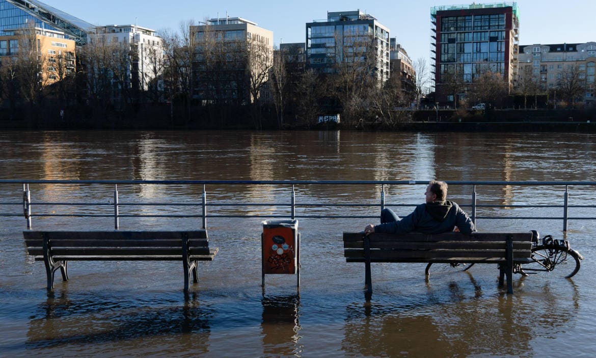Person sitzt auf einer Bank am überschwemmten Flussufer bei Hochwasser.
