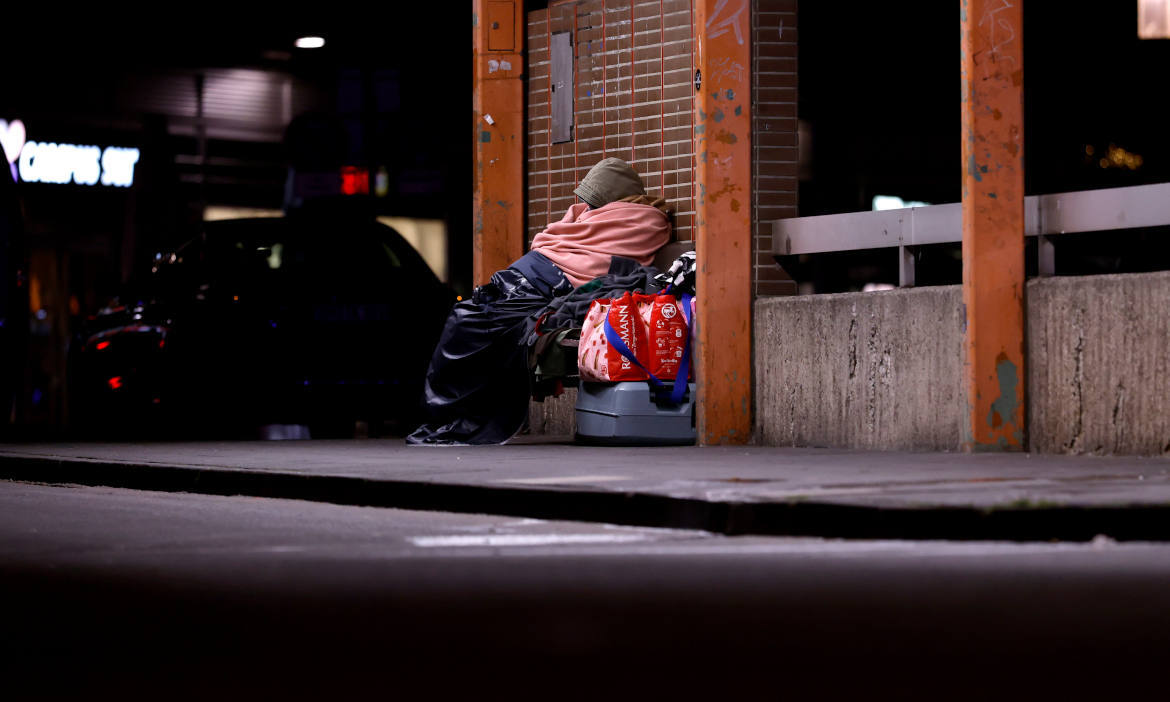 Obdachlose Person sitzt an einer Bushaltestelle mit Gepäck und Decke, symbolisiert Armut und soziale.