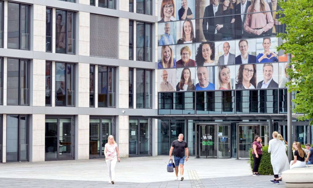 Modernes Bürogebäude mit großem Portrait-Fenster, Menschen vor dem Eingang.