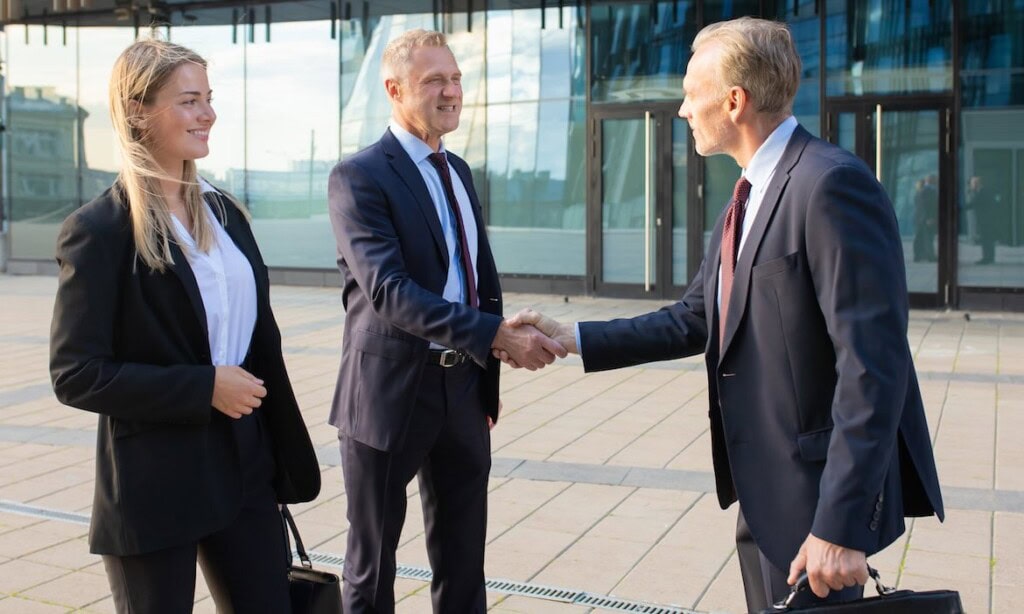 Zwei Geschäftsleute beim Händeschütteln vor moderner Firmenfassade.