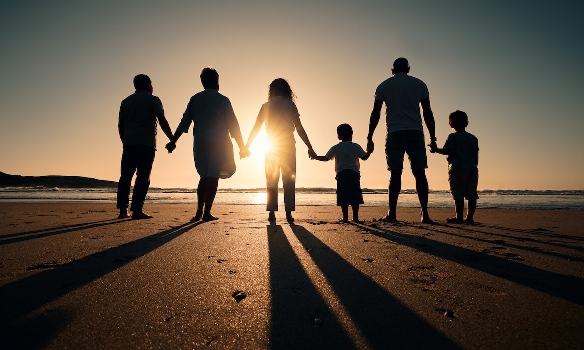 Familie hält Händchen am Strand bei Sonnenuntergang, symbolisiert Zusammenhalt.