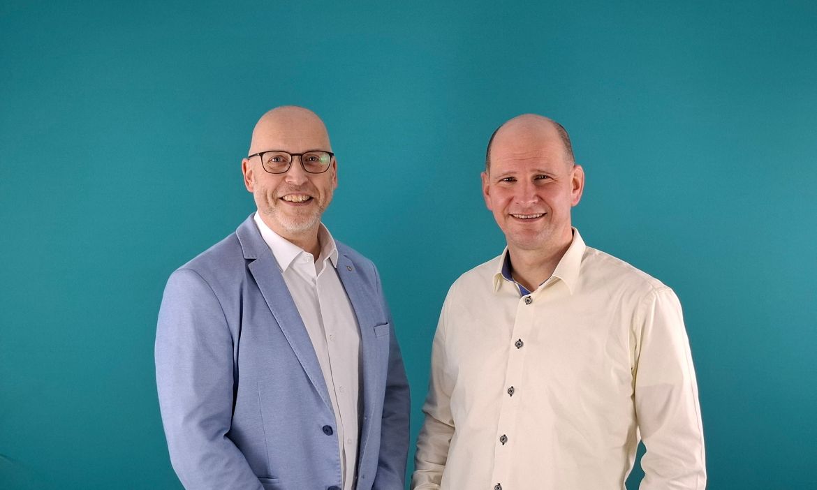 Zwei Männer in Business-Kleidung vor einem blauen Hintergrund.