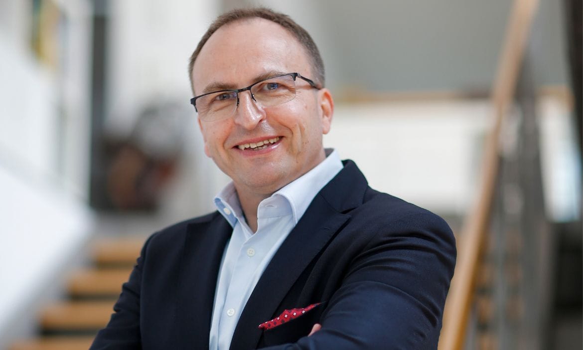 Mann im Business-Outfit mit Brille, lächelnd, in einem modernen Büro.