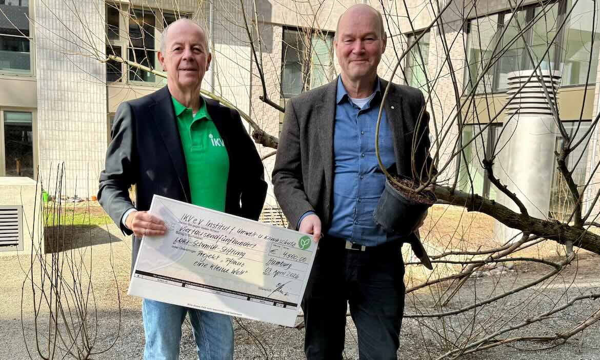 Zwei Männer präsentieren eine Spendenurkunde vor einem Baum im Schulhof.