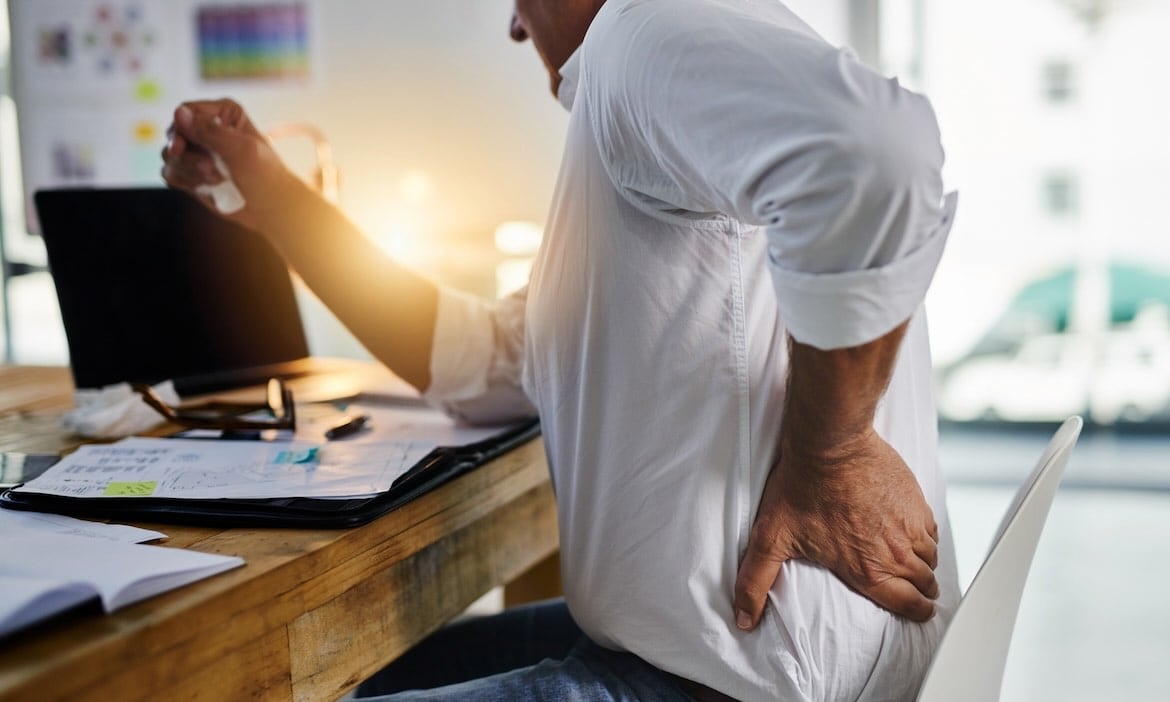 Mann mit Rückenschmerzen bei der Arbeit im Büro, hält sich den unteren Rücken.