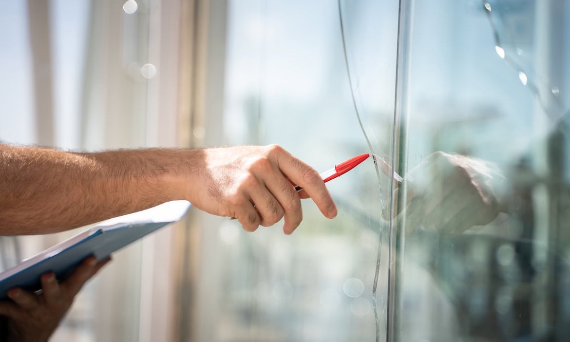 Hand, die mit einem roten Marker eine Glasscheibe markiert, im Hintergrund Fenster.