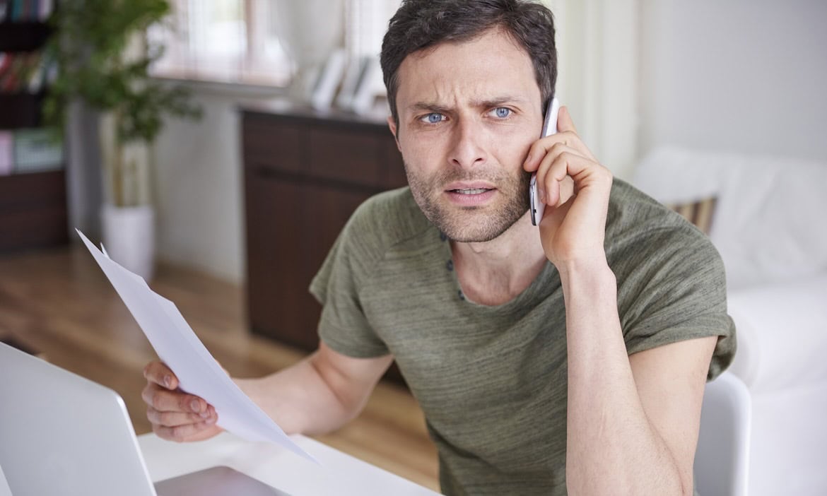 Mann am Telefon mit besorgtem Gesicht, hält Papier in der Hand, im Hintergrund ein modernes Wohnzimm.