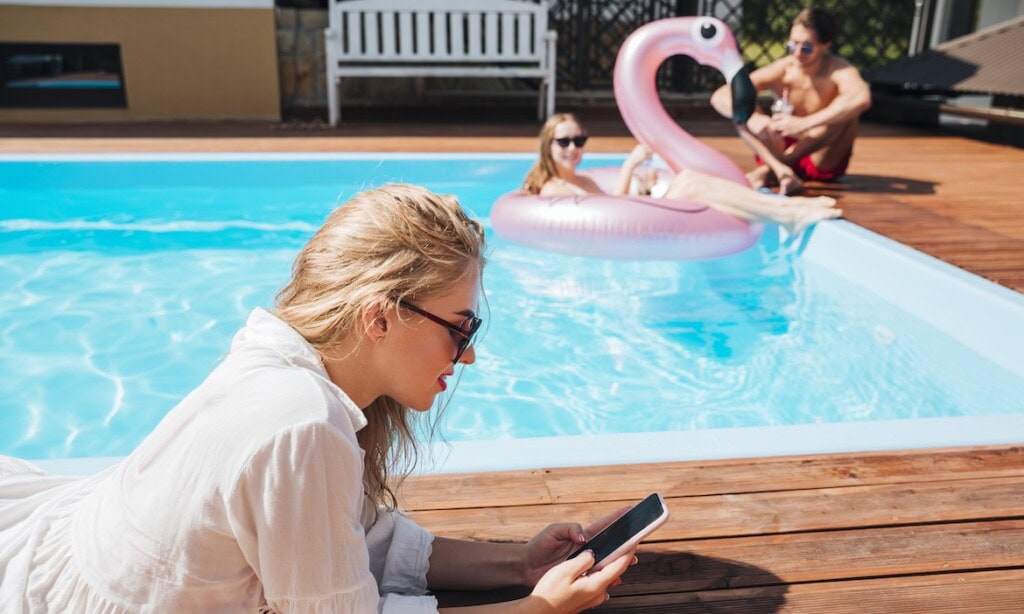 Frau liest am Pool, Kinder mit Flamingo-Inflatable im Wasser, sonniger Tag.