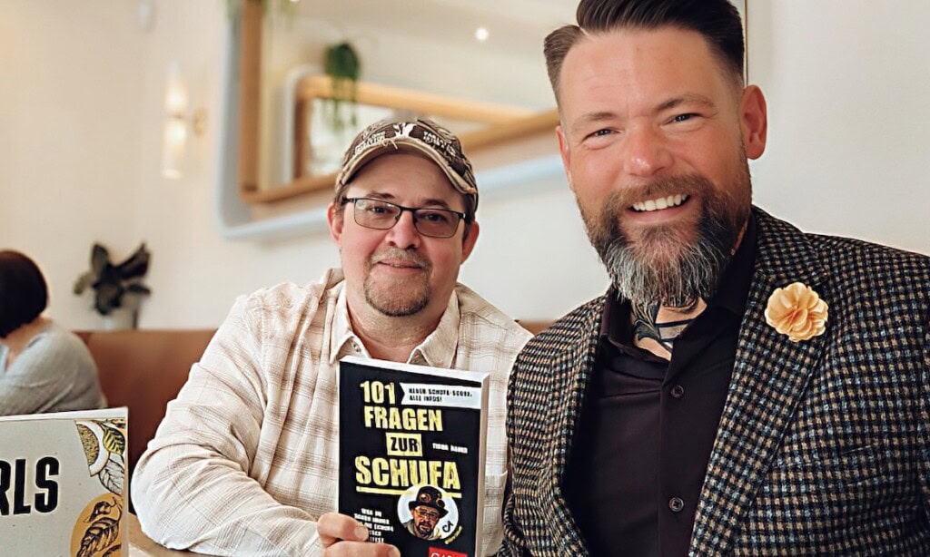 Zwei Männer diskutieren über Schufa im Café, einer hält ein Buch in der Hand.