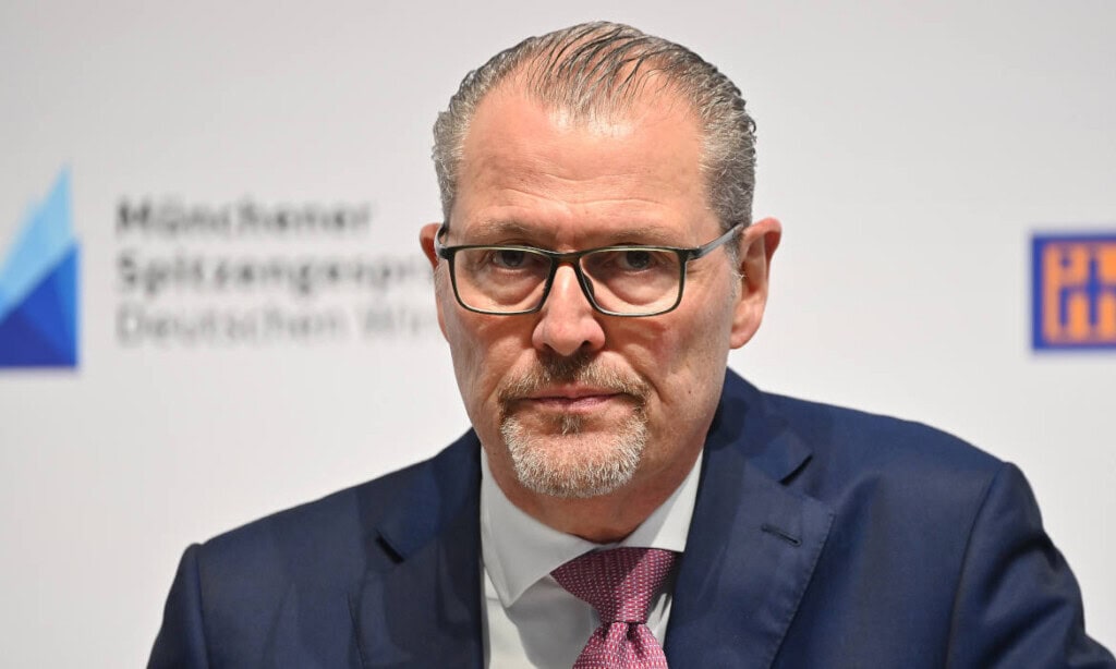Der Mann in Anzug bei einer Konferenz, professionell gekleidet, mit Brille und grauem Haar.