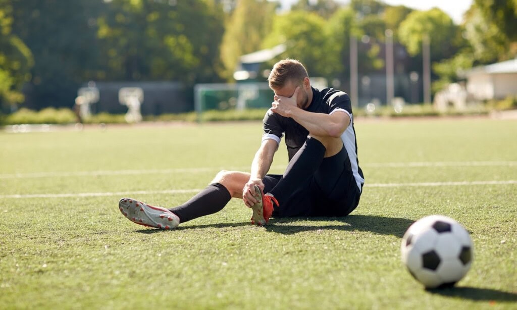 Fußballspieler sitzt erschöpft auf dem Spielfeld, hält sich den Knöchel, nach einem Spiel.