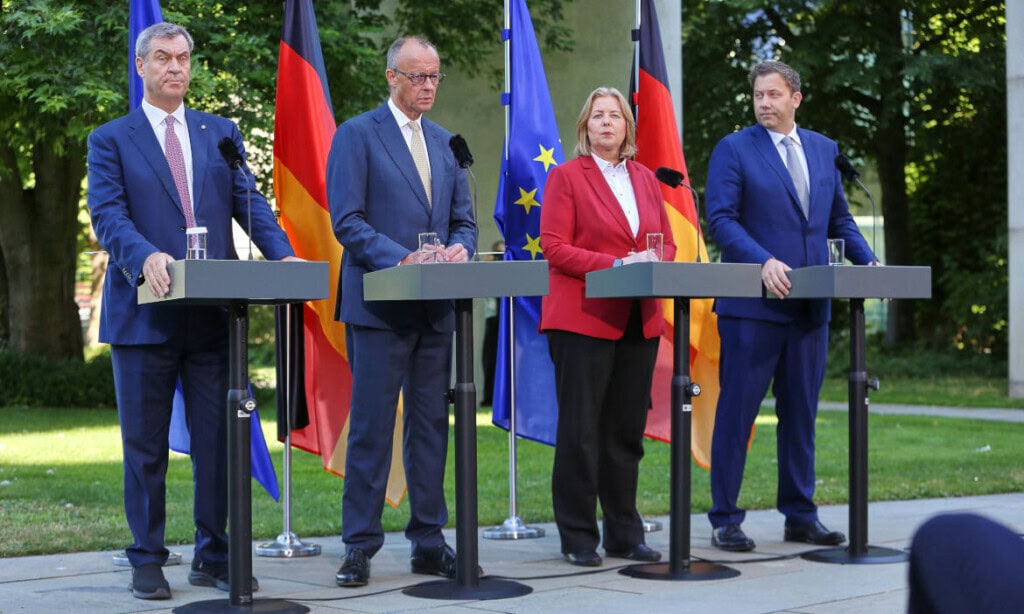 Politiker bei Pressekonferenz mit Deutschland- und EU-Flaggen im Hintergrund.