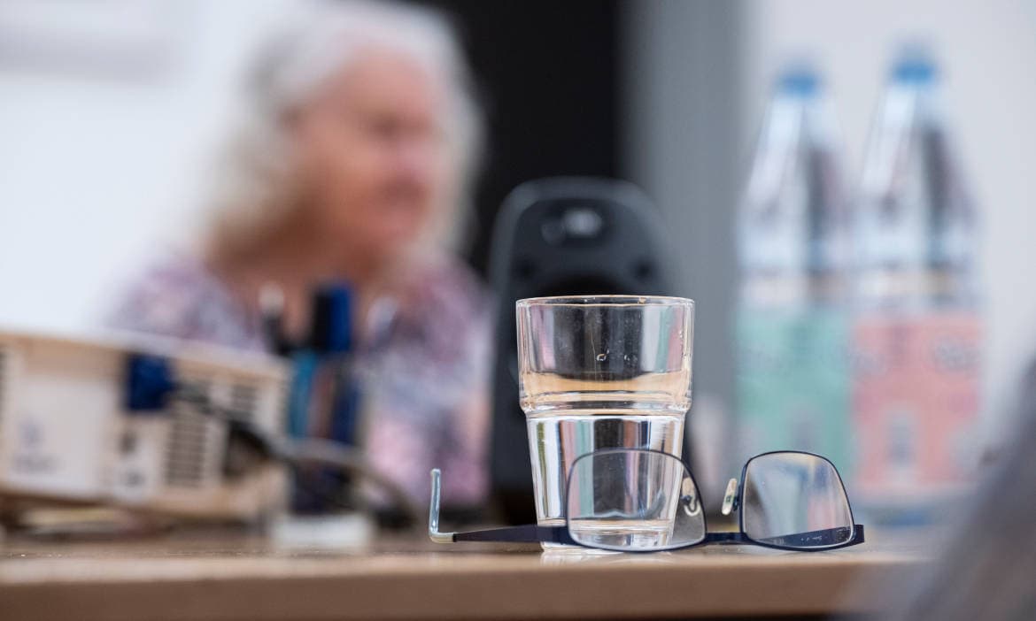 Glas Wasser, Brille und Smartphone auf Tisch bei Pflegekraft im Hintergrund.