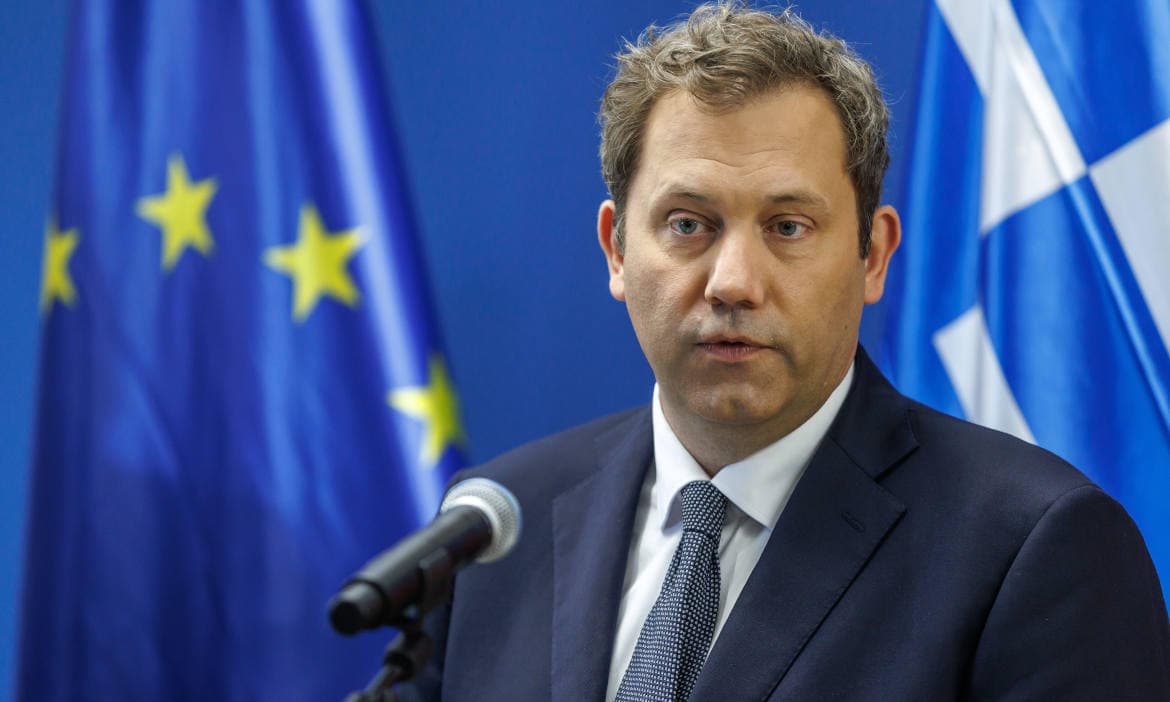 Mann bei einer Pressekonferenz vor EU- und griechischer Flagge.