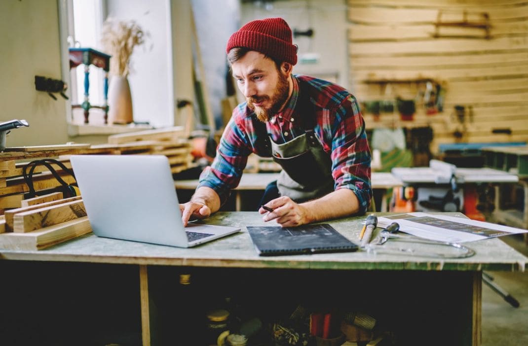 Handwerker arbeitet an Laptop in Werkstatt mit Werkzeugen und Holzmaterialien.