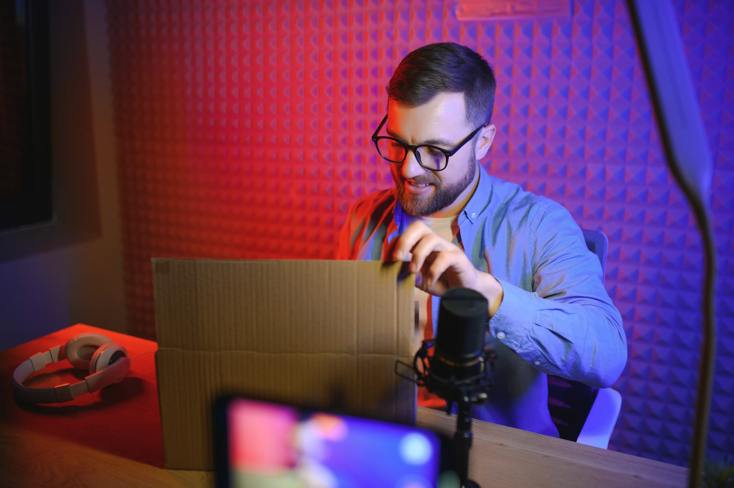 Junger Mann öffnet Paket in Tonstudio mit Mikrofon und Kopfhörern.